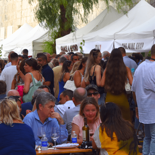 A lively outdoor wine festival in Gibraltar with many people gathered around white tents and tables, some sitting and enjoying wine, and others standing and socializing.