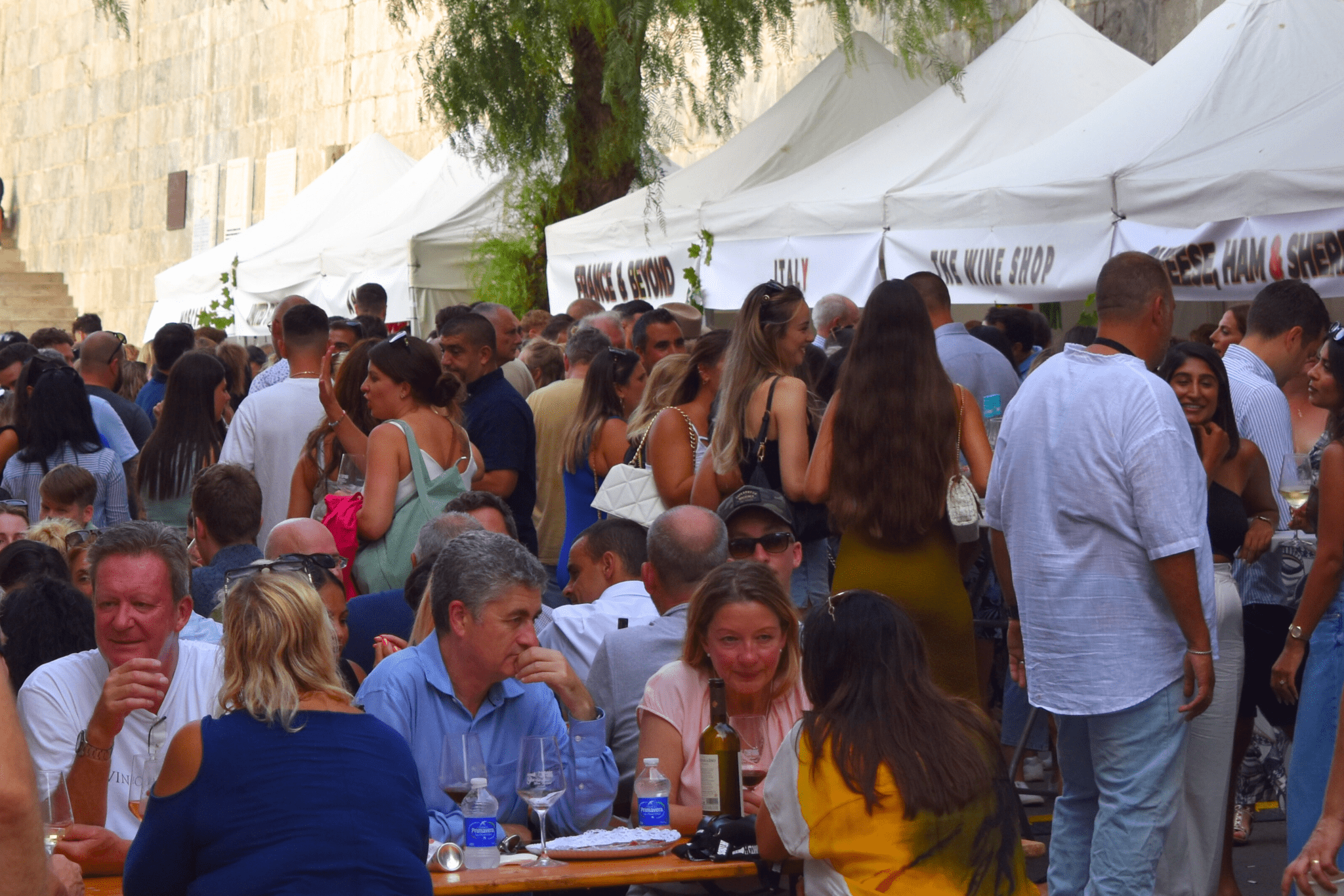 A lively outdoor wine festival in Gibraltar with many people gathered around white tents and tables, some sitting and enjoying wine, and others standing and socializing.